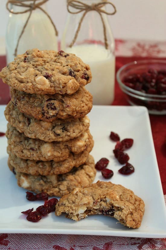 Oatmeal Craisin White Chocolate Chip Cookies stacked up on a white plate with craisins on the plate and a glass bowl of craisins in the background.