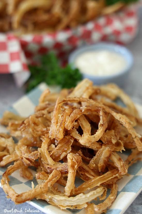 Fried Onion Strings are the perfect snack recipe for any day. Friend Onion Strings on a blue and white checkered plate with small blue bowl filled with ranch, along with parsley in the background.