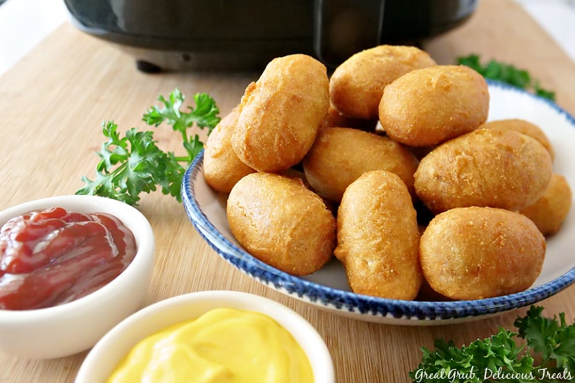 Mini Corn Dogs in Air Fryer are super quick and easy to make, and are super crunchy and crispy. a white plate with blue trim loaded with mini corn dogs, a small bowl full of ketchup and another small bowl of mustard.