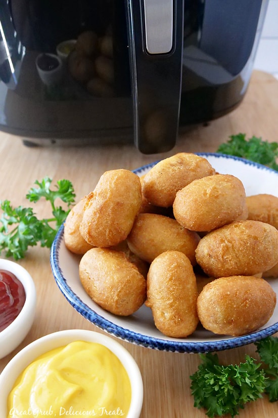 Mini Corn Dogs in Air Fryer is a super simple and easy air fryer recipe to make that gives you crispy corn dogs in a matter of minutes. A plate of mini corn dogs with ketchup next to it, and the air fryer in the background.