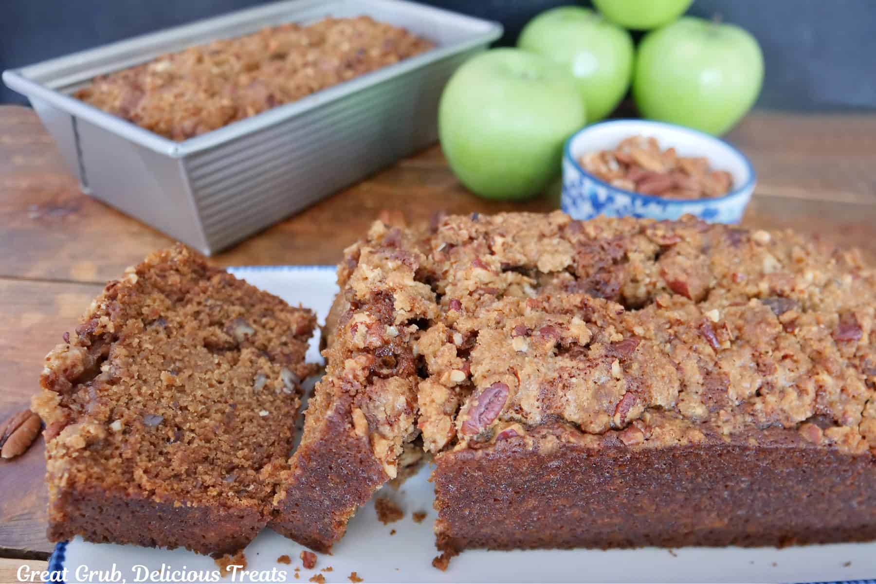 A wood surface with a white plate with blue trim and a silver loaf pan both with apple bread on them.