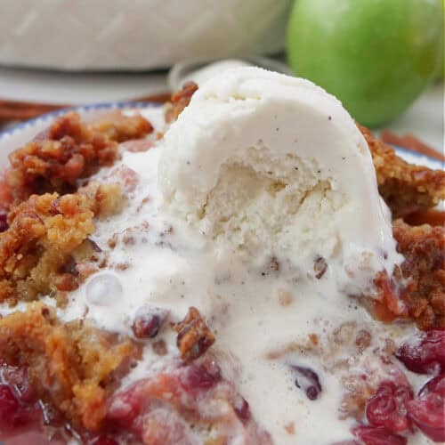 A white bowl with blue trim with a serving of apple cranberry crisp topped with vanilla ice cream which has started to melt.