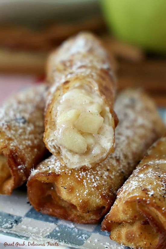 Apple Cream Cheese Egg Rolls are filled with a homemade apple cream cheese filling for a crispy, delicious dessert. Four fried apple egg rolls on a blue and white checkered plate with a bite taken out of one of them.
