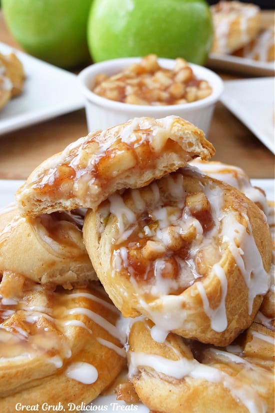 A half a dozen apple pinwheels stacked up on a white plate with a bite taken out of the pinwheel on top.