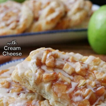 Two apple pastries on a white plate with blue trim with a bite taken out of one of the pastries with more pastries in the background.