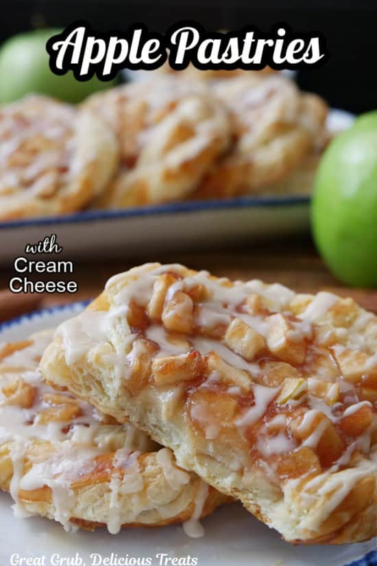 Two apple pastries on a white plate with blue trim with a bite taken out of one of the pastries with more pastries in the background.