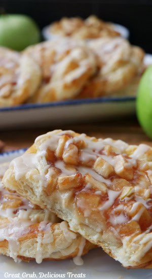 Two apple pastries on a white plate with more pastries in the background.