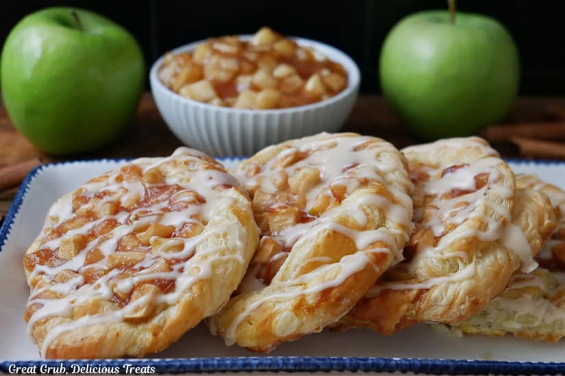 A horizontal photo of four apple pastries on an oblong white plate with blue trim, and a small white bowl filled with apple filling, and two green apples in the background.