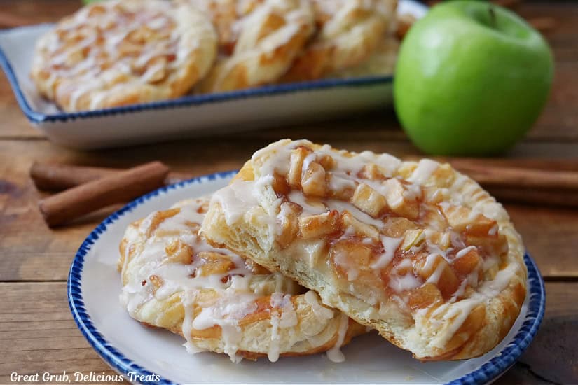 A horizontal photo of a wood surface with a round white plate with blue trim with two apple pastries on it with a bite taken out of one of the pastries, and more pastries in the background.
