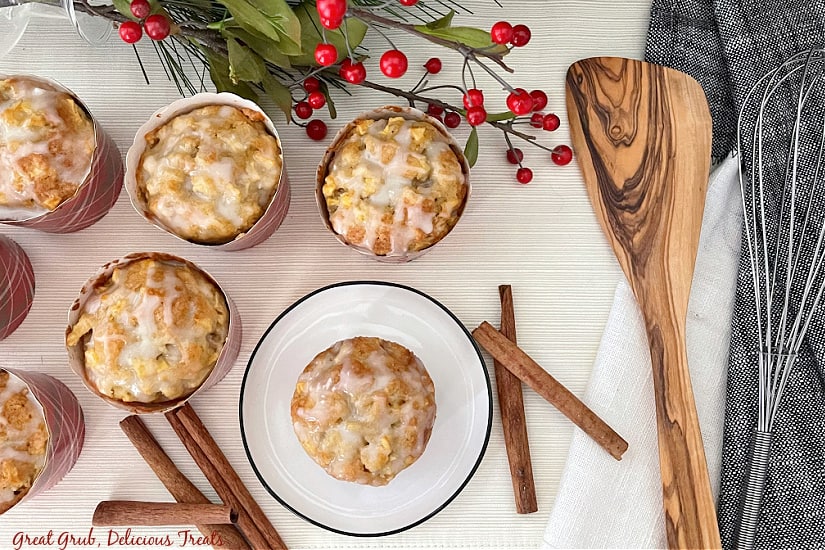 Apple Muffins with Glaze are a delicious homemade muffin recipe that is loaded with diced apples and topped with a homemade vanilla glaze. An overhead photo of apple muffins sitting on a white background, with cinnamon sticks, a whisk, a wooden spoon, and apple muffins with vanilla glaze.