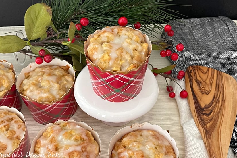 Apple Muffins with Glaze are soft, moist, and full of fresh diced apples and topped with a simple vanilla glaze. Apple muffins in red plaid cupcake liners, all topped with a glaze, with one muffin sitting on a white cupcake stand.
