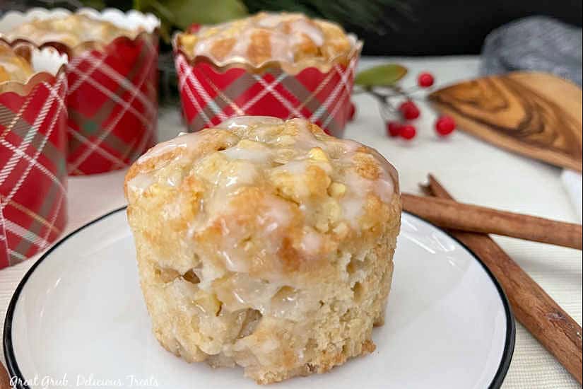 Apple Muffins with Glaze are a great recipe to whip up for a dessert or for a delicious breakfast. A muffin sitting on a white plate with black trim, with cinnamon sticks and apple muffins in the background.