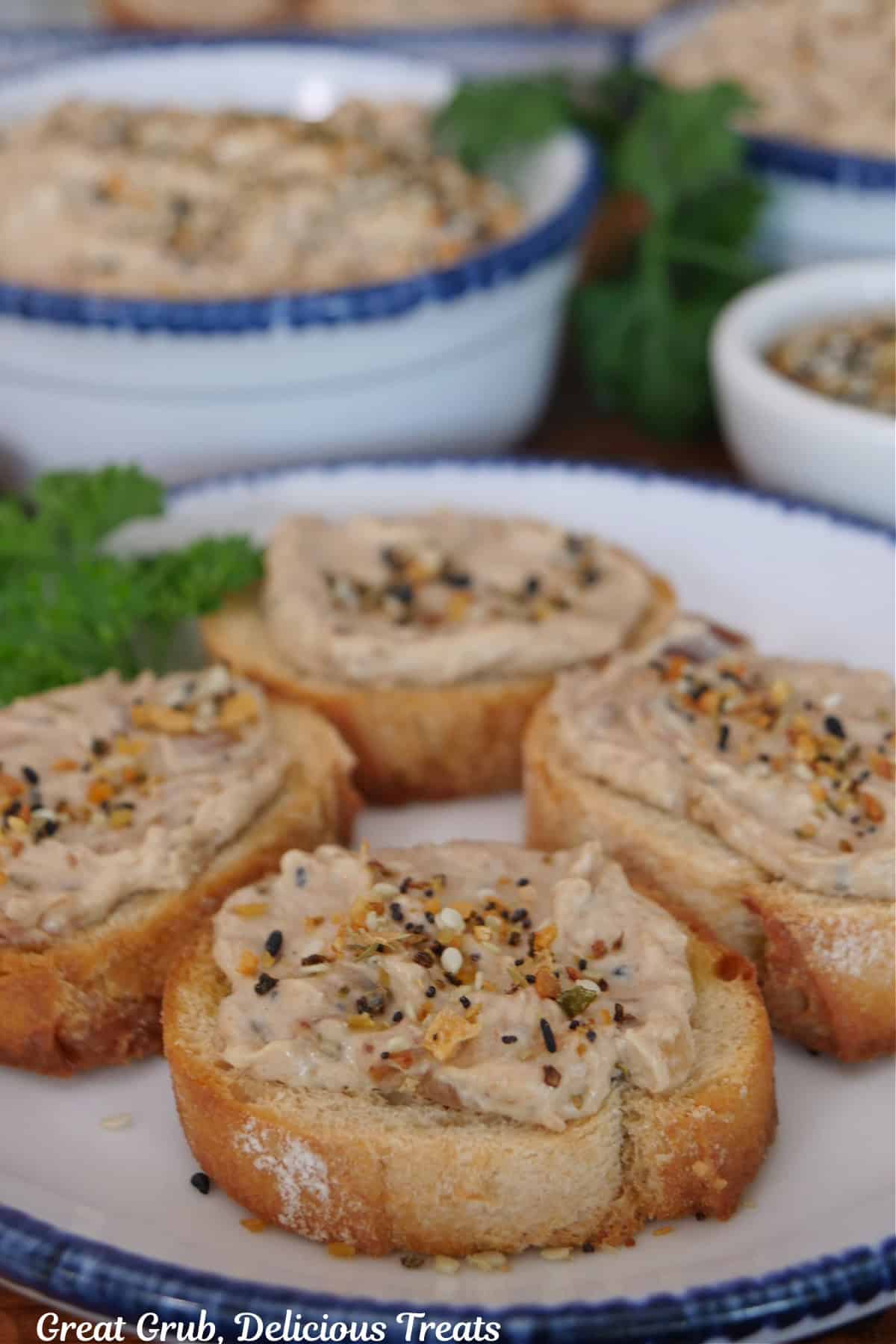 A wood surface with white bowls with blue trim with slices of baguettes topped with bacon jam on it and two bowls filled with the bacon jam in the background.