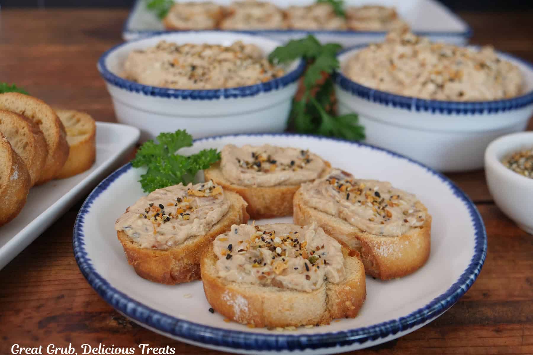 A wood surface with white bowls with blue trim with slices of baguettes topped with bacon jam on it and two bowls filled with the bacon jam in the background.