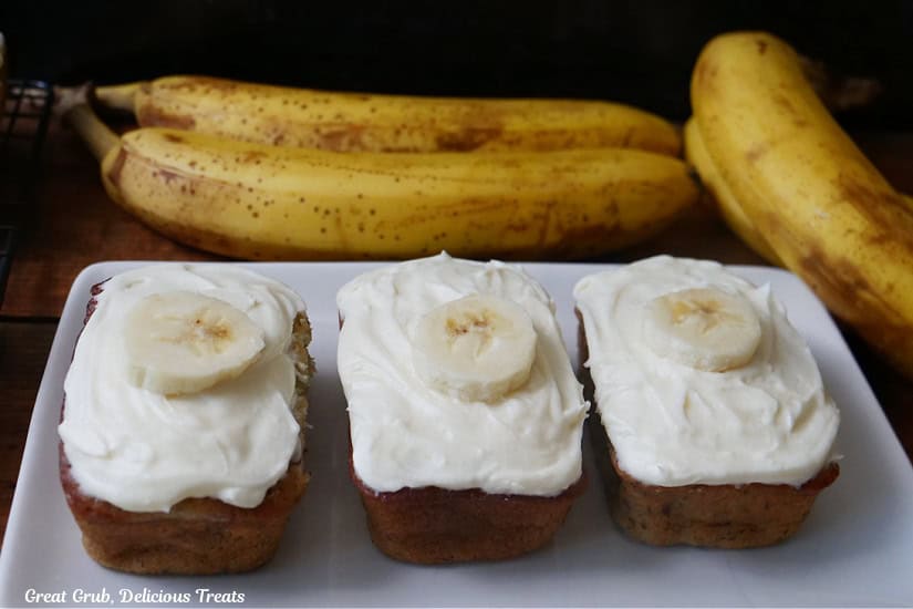 A white oblong plate with three mini loaves of banana nut bread with cream cheese frosting on them with ripe bananas in the background.