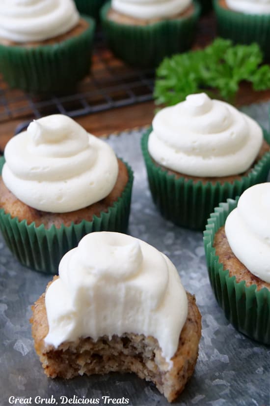 A few banana nut muffins with cream cheese frosting on top placed on a silver tray and more in the background, and one with a bite taken out of it.