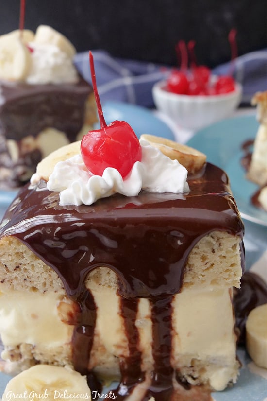 A square slice of banana cake with hot fudge on the top with whipped cream and a cherry.
