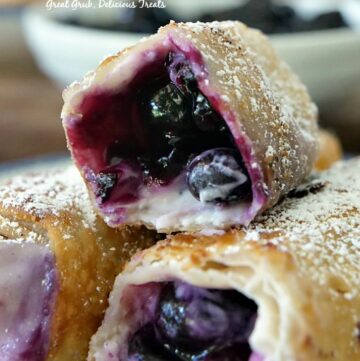 Three blueberry egg rolls on a white plate with blue trim showing the filling inside.