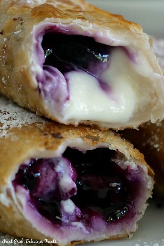 Blueberry Cream Cheese Egg Rolls are filled with cream cheese and fresh blueberries, then fried and sprinkled with powdered sugar. A close up photo of two egg rolls with bites taken out of them showing the inside filling.