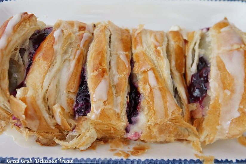 A white plate with blue trim with a blueberry pastry braid on it that has been cut into slices.
