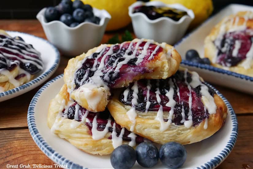 A horizontal photo of three white plates with blue trim with blueberry danishes on them.