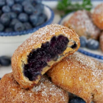 A white plate with three fried blueberry pie bombs with a bite taken out of the one on top with the title of the recipe at the top.