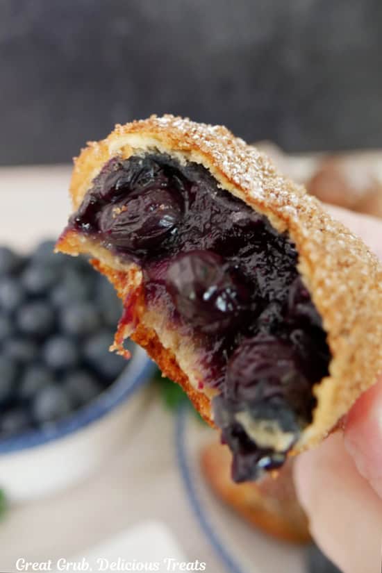 A blueberry pie bomb held up with a bite taken out of it showing the blueberry filling inside.