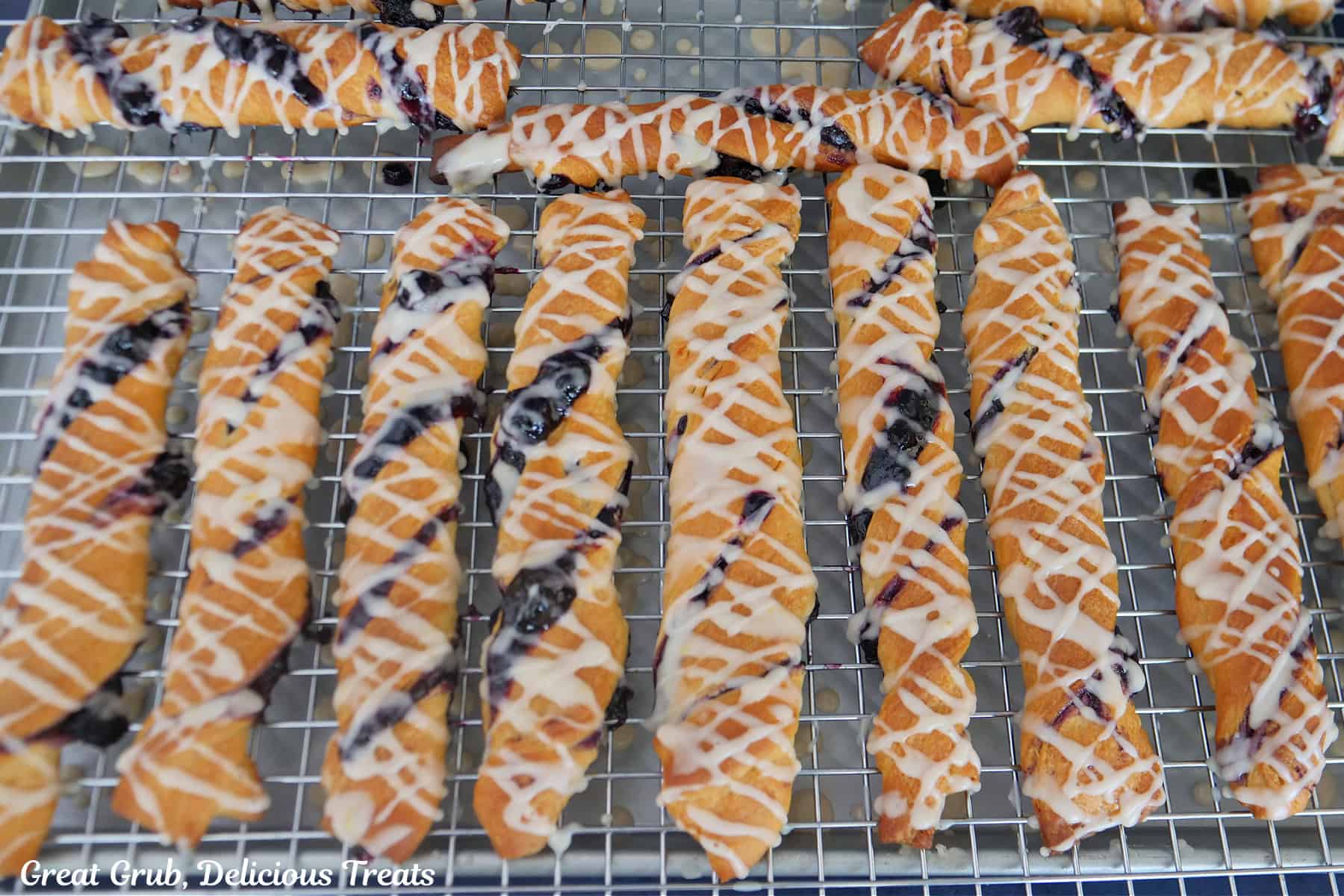 A baking sheet with a wire rack on top with 14 blueberry twists on them that have been drizzled with lemon glaze.