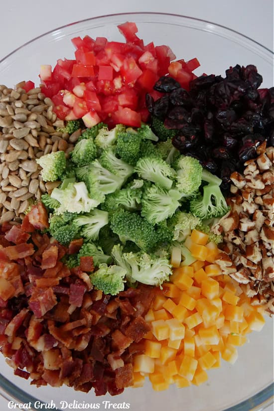 A clear glass bowl filled with chopped broccoli, bacon, pecans, tomatoes, craisins, and sunflower seeds.