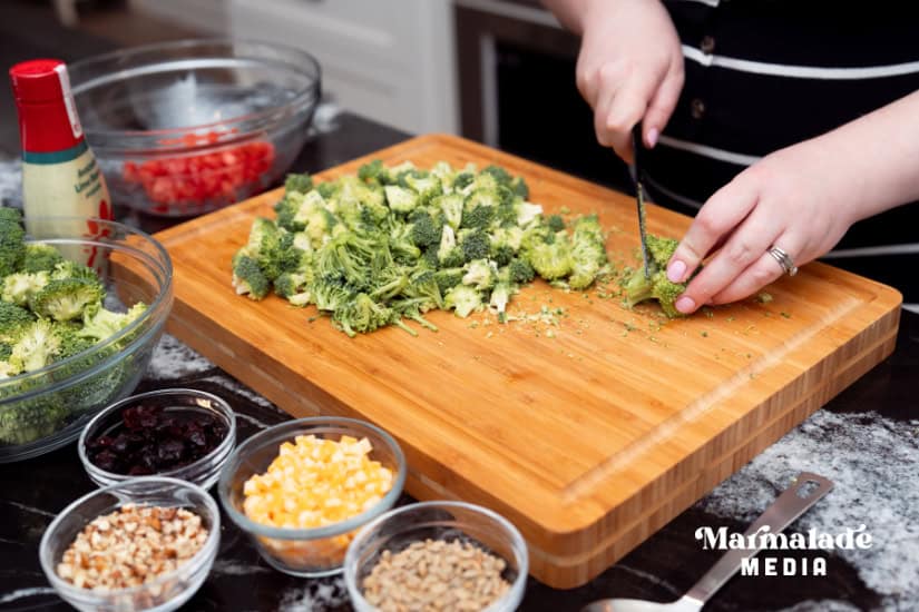 A cutting board with broccoli florets being chopped and the other ingredients in clear glass bowls.