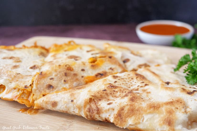 A horizontal photo of a wood cutting board with a buffalo chicken quesadilla on it that has been cut into three pieces.