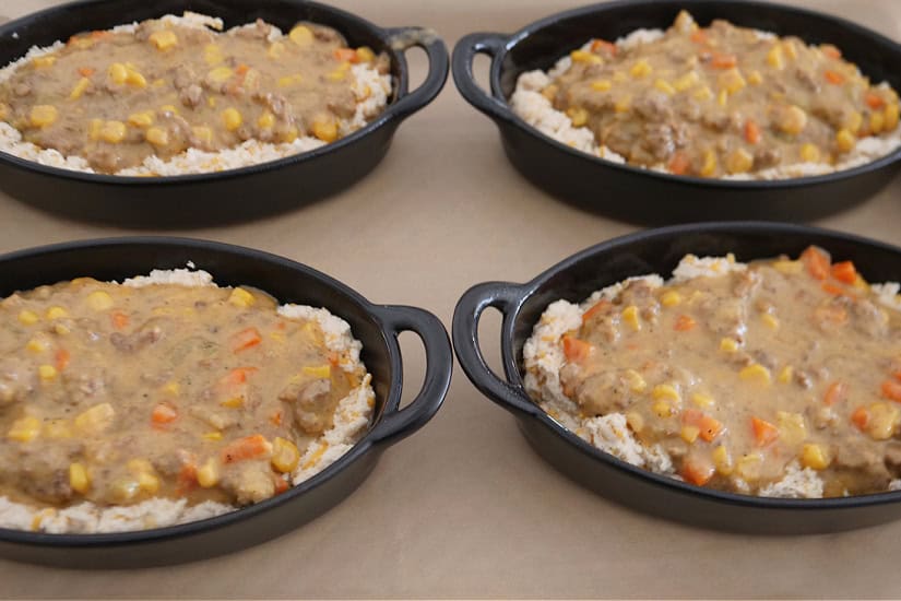 A baking sheet with four oval black baking dishes filled with the meat mixture over the biscuit bottom.
