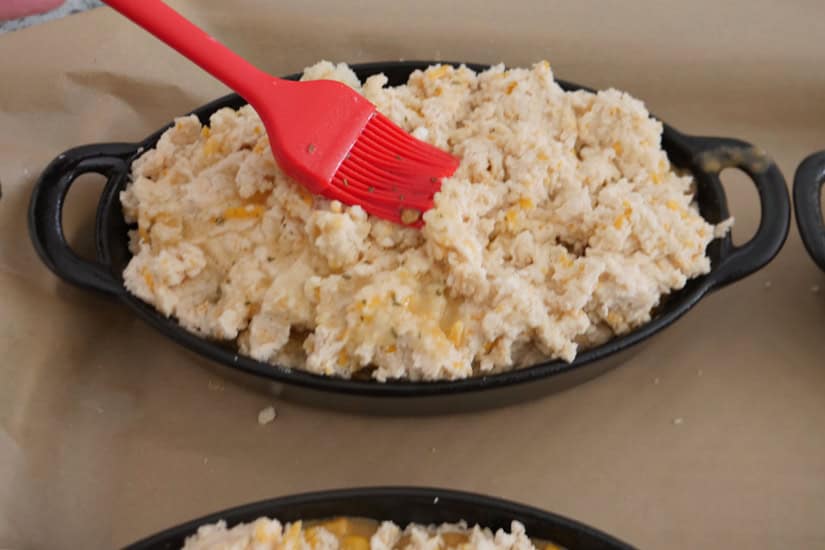 A red silicone brush brushing melted butter over the top of a cheddar bay beef pot pie.