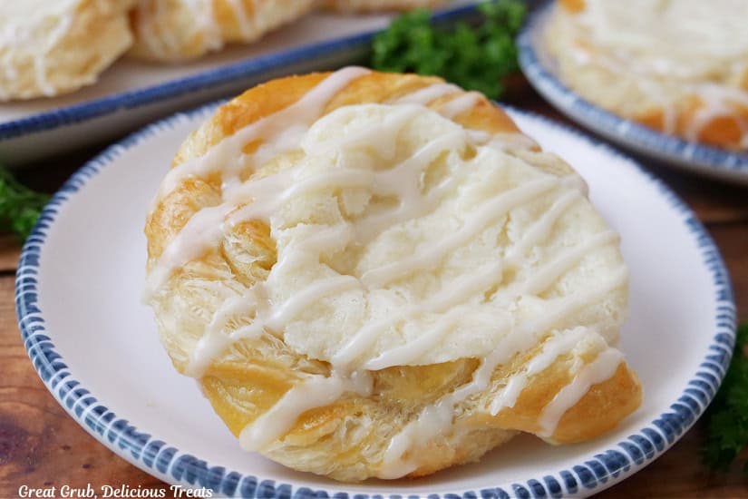 A white plate with blue trim with a cheese danish on it with other danishes in the background.