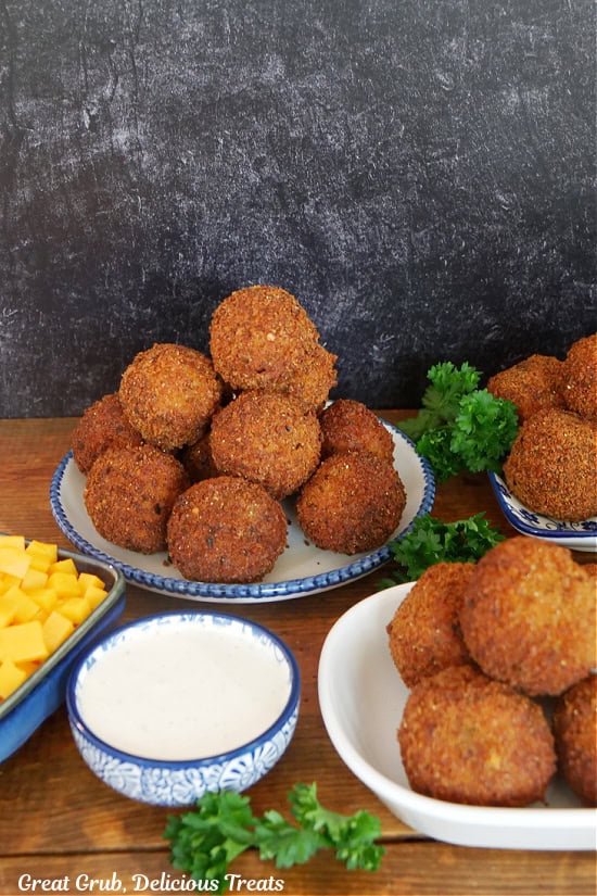 A wood surface with white plates with blue trim filled with cheesy ground beef balls on it with a small bowl of ranch dressing and diced cheese.
