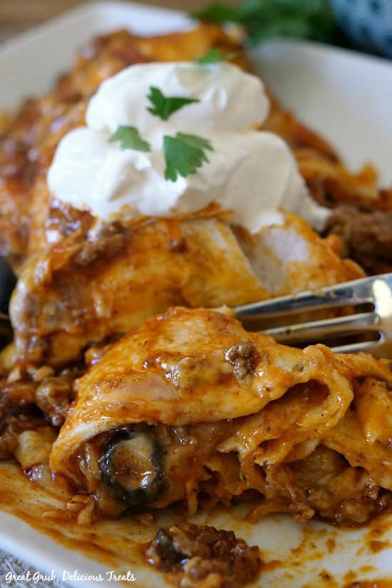 Cheesy Ground Beef Enchiladas are delicious and hearty, full of ground beef, two types of cheese and topped with sour cream. A white plate with an enchilada on it and a fork cutting a bite off the enchilada.