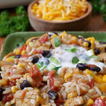 A square green plate with a serving of chicken enchilada casserole, rice, beans, corn with sour cream on top.