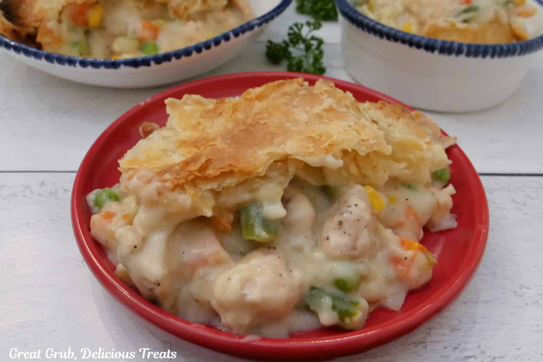 A white surface with a red plate and two white plates with a serving of chicken pot pie casserole on each.