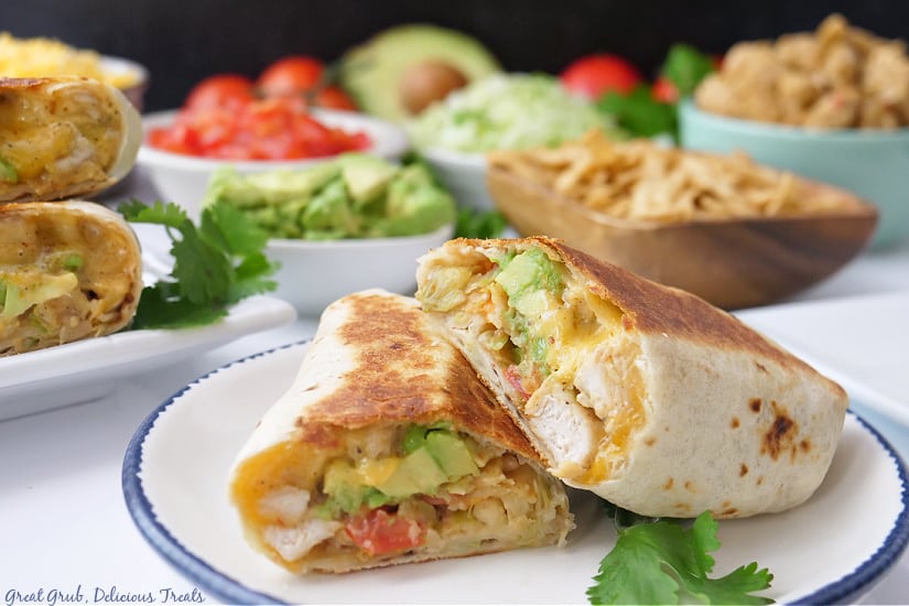 A white plate with blue trim with a grilled chicken burrito on it that is cut in half with all the different ingredients in the background.