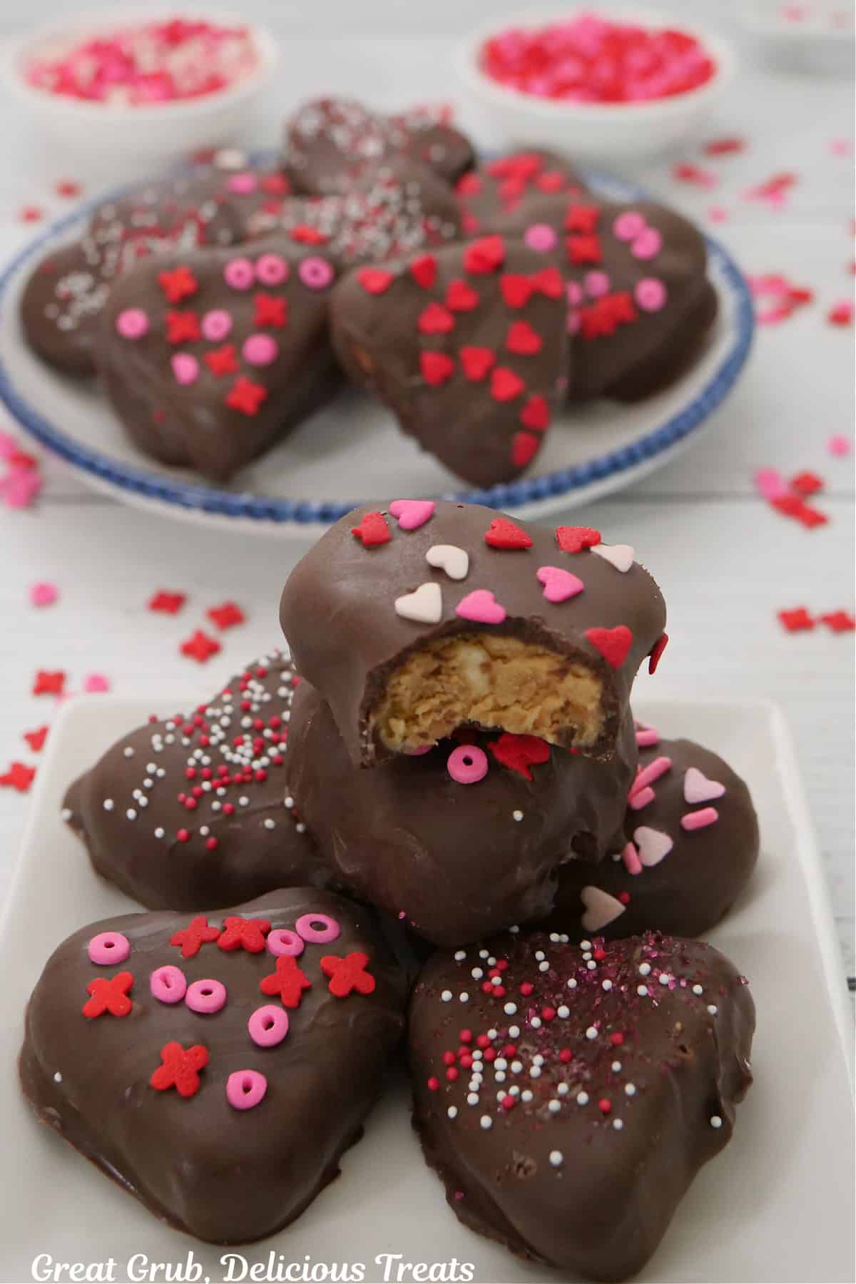 A white surface with 14 chocolate covered hearts on it with candy sprinkles scattered around.