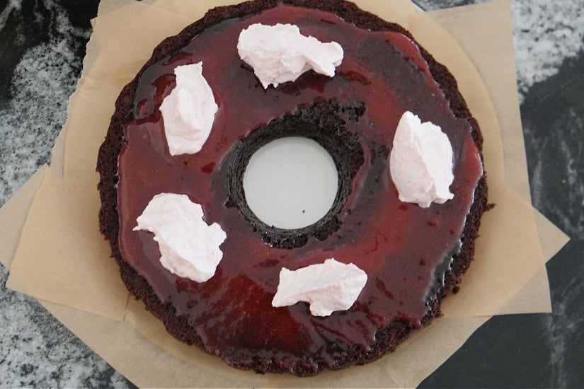 A black surface with the bottom half of a chocolate bundt cake on it with a strawberry filling and strawberry cream frosting on it.