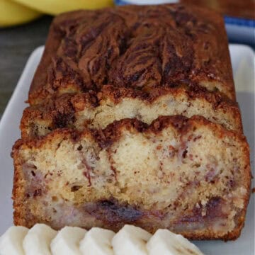 A white plate with a loaf of banana bread on it that has been sliced with sliced bananas on the plate.