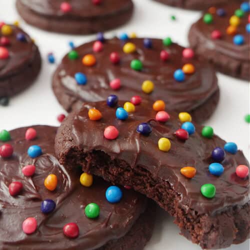 A white surface with cosmic brownie cookies on it with rainbow sprinkles scattered around the cookies.