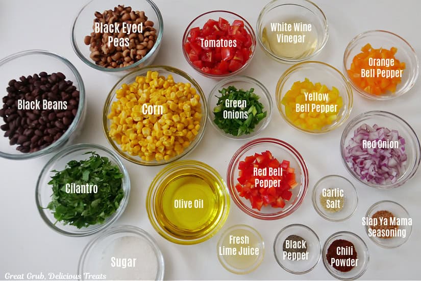 A white surface with all of the ingredients for cowboy caviar in glass bowls with the title of each ingredient typed on top of each ingredient.