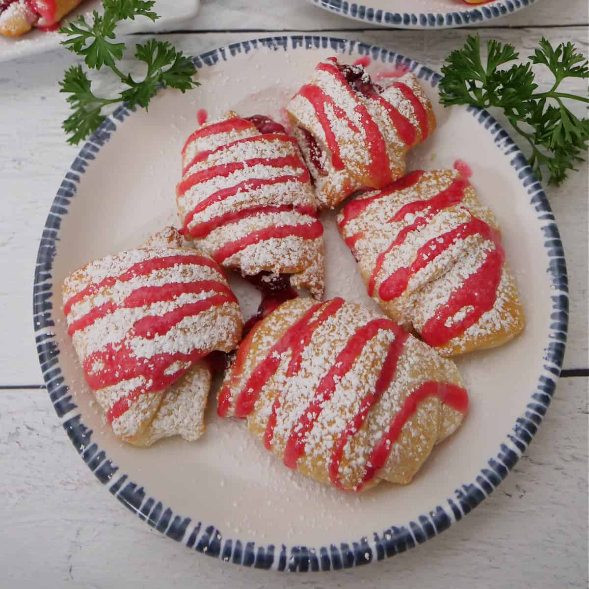 A white surface with a round white plate with blue trim with five cranberry cream cheese pastries on it.