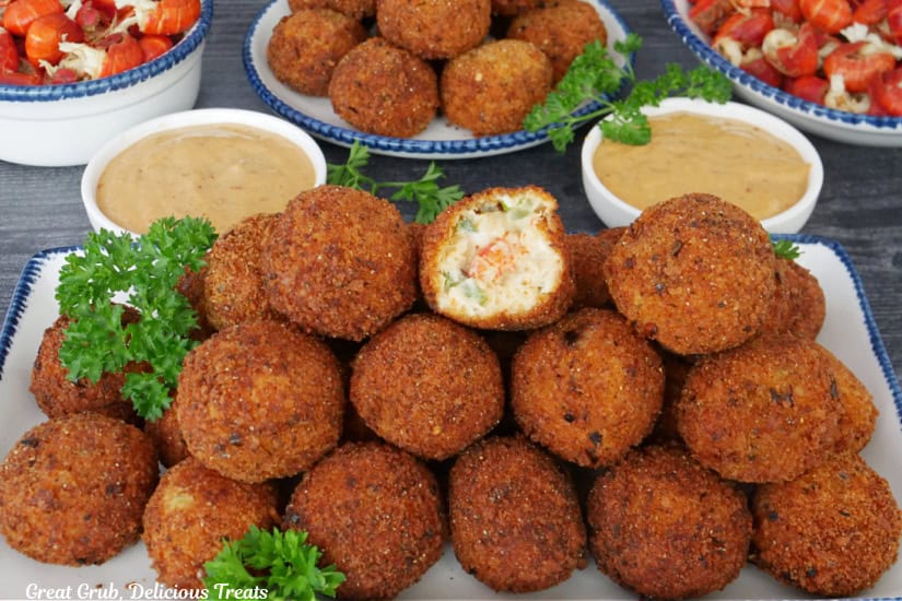 An oblong white plate with blue trim with about 20 crawfish balls on it with two bowls filled with crawfish tails and a round plate with more crawfish balls on it in the background.
