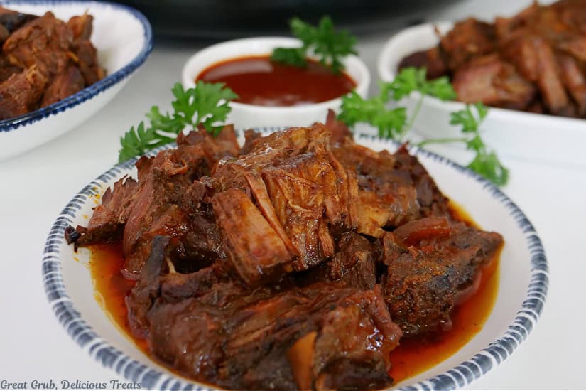 A white round plate with blue trim with boneless beef ribs on it with a small white bowl filled with BBQ sauce in the background.