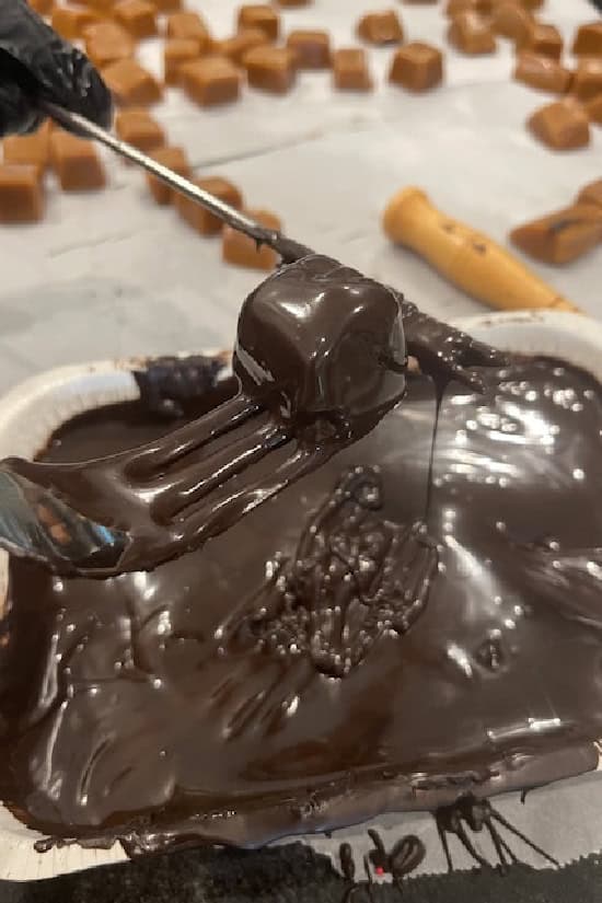 A piece of caramel being dipped and covered in dark chocolate.