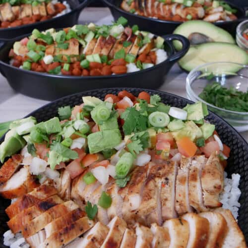 A white surface with grilled chicken, rice, avocado, pico in black bowls.