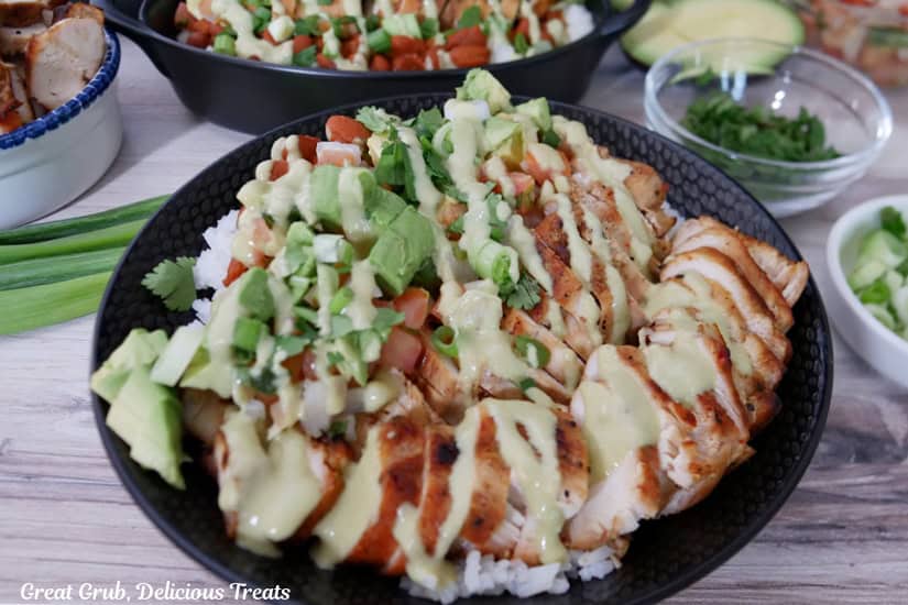 A black bowl on a light colored surface with all the ingredients of grilled chicken burrito bowls in it with other black bowls in the background.